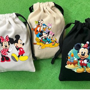 May include: Three drawstring bags with different Disney character designs. The first bag is white with Mickey Mouse and Minnie Mouse. The second bag is beige with Mickey Mouse, Minnie Mouse, Donald Duck, Goofy, and Pluto. The third bag is black with Mickey Mouse wearing a birthday hat.