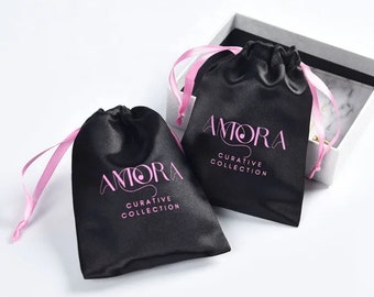 Könnte beinhalten: Zwei schwarze Satin-Säckchen mit rosa Kordelzugbändern. Beide Taschen haben den Text "AMORA CURATIVE COLLECTION" in rosa gedruckt.