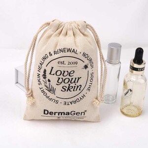 Op de afbeelding: Een natuurlijke beige trekkoordtas met de tekst "Love your skin" in een cirkel met een bloemenontwerp. De tas is gelabeld "DermaGen" en heeft de tekst "est. 2019" en "Skin Healing & Renewal Nourish Soothe Hydrate" rond de cirkel. De tas ligt naast twee heldere glazen flessen met druppelaars.