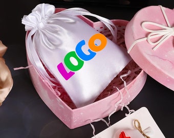 Könnte beinhalten: Eine weiße Satin-Kordeltasche mit dem Wort "LOGO" in bunten Buchstaben bedruckt. Die Tasche befindet sich in einer rosa herzförmigen Schachtel mit weißem Seidenpapier.