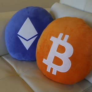 May include: Two plush pillows, one blue with a white Ethereum logo and one orange with a white Bitcoin logo.