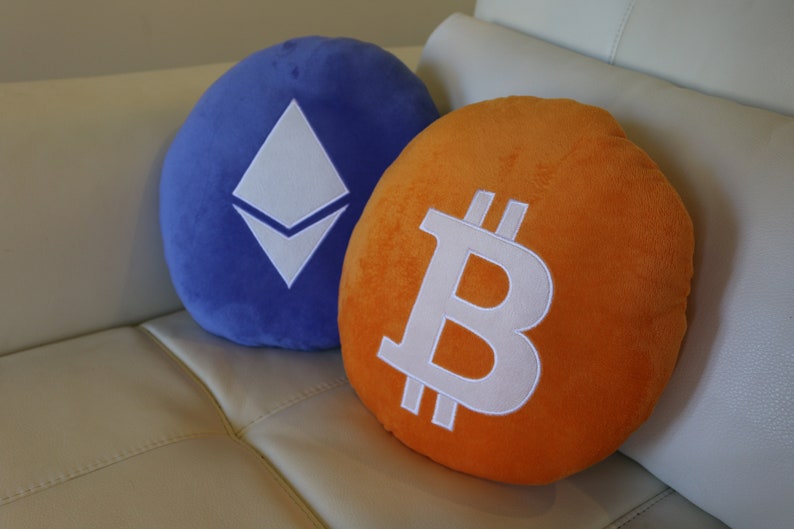 May include: Two plush pillows, one blue with a white Ethereum logo and one orange with a white Bitcoin logo.