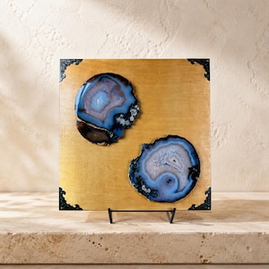 May include: A square gold-toned panel featuring two circular blue and white agate slices mounted on the surface. The panel has decorative black metal corner accents and is supported by a black metal stand, creating a display piece.