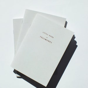 Può includere: Una pila di tre piccoli quaderni bianco sporco. Il quaderno superiore presenta la scritta "LITTLE BOOK of PASSWORDS" in un carattere marrone scuro. I quaderni sono su una superficie bianca, con un'ombra scura.