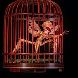 Op de afbeelding: Een gedetailleerde, handgemaakte houten vogelkooi met een roodbruine afwerking. Binnenin is een grotesk, gevleugeld wezen met zichtbare tanden en klauwen te zien. De kooi staat op een donkere ondergrond, met een klein stukje groen gras aan de basis.
