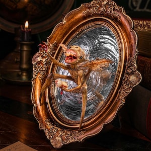 Puede incluir: Espejo ovalado antiguo con marco dorado ornamentado. Dentro del espejo, una criatura grotesca y esquelética con alas y dientes afilados sostiene un pequeño objeto. El fondo incluye libros y una vela encendida, creando un ambiente vintage.