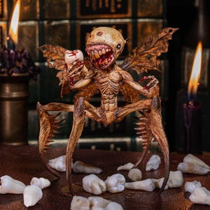 Puede incluir: Figura detallada de una criatura fantástica marrón con patas y alas de insecto, sosteniendo un diente. La criatura tiene una amplia sonrisa dentuda y está rodeada de varios dientes sueltos. Velas encendidas y libros antiguos en el fondo.