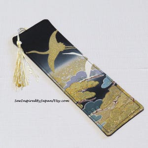 May include: A rectangular bookmark with a black background and a gold crane design. The bookmark features a gold tassel and a pattern of clouds and trees in gold, blue, and green. The text "SewInspiredByJapan/Etsy.com" is visible.