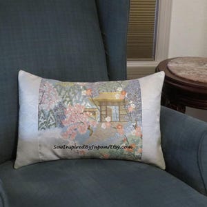 May include: Rectangular decorative pillow with a Japanese-inspired design. The center features a landscape with a building, cherry blossoms, and a light blue background. The sides are a light cream color. The pillow is placed on a teal armchair.