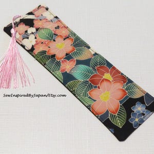 May include: A black fabric bookmark with a pink tassel. The bookmark features a floral pattern with pink, red, and green flowers on a black background. The text "SewInspiredByJapan/Etsy.com" is printed on the bookmark.