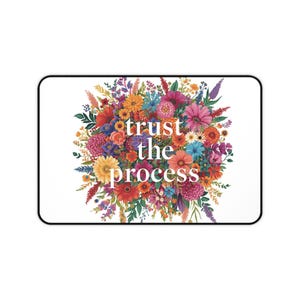 Puede incluir: Alfombrilla de ratón rectangular con borde negro que presenta un vibrante diseño floral. El diseño incluye una variedad de flores y follaje coloridos, con las palabras "trust the process" en el centro. El fondo es blanco.