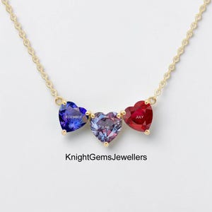 May include: A gold-colored necklace featuring three heart-shaped gemstones in blue, purple, and red. Each gemstone has a month name inscribed on it. The necklace is from KnightGemsJewellers.