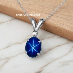 Puede incluir: Un collar de plata con un colgante que presenta una piedra preciosa de zafiro azul estrella con una estrella de seis puntas. El colgante está engastado en un marco ovalado de plata y tiene un pequeño acento de diamante sobre el zafiro.
