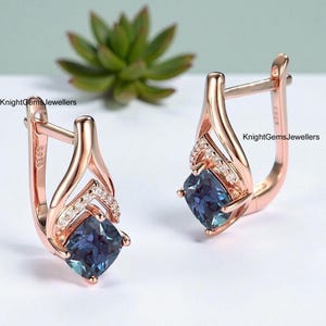 Op de afbeelding: Roségouden oorbellen met een vierkante, donkerblauwe edelsteen en kleine witte accentstenen. De oorbellen hebben een klapsluiting. De tekst "KnightGemsJewellers" is zichtbaar.