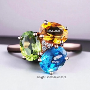 May include: A rose gold ring with three oval gemstones in green, yellow, and blue, with the months "AUGUST", "NOVEMBER", and "MARCH" inscribed. Small diamonds are set in the centre of the ring.