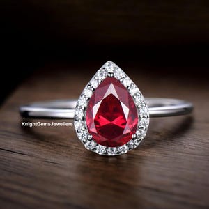 Può includere: Un anello color argento con una pietra preziosa rossa a forma di pera al centro, circondata da un alone di piccole pietre bianche. La fascia è sottile e metallica. Il testo KnightGemsJewellers è visibile sul lato sinistro della montatura.