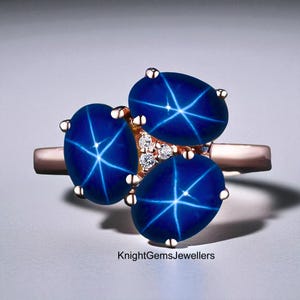 Puede incluir: Un anillo de oro rosa con tres zafiros estrella ovalados de color azul oscuro. Los zafiros tienen un efecto de estrella y están engastados con pequeños diamantes. El anillo es de KnightGemsJewellers.