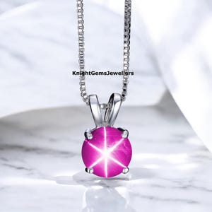 Peut inclure: Un collier pendentif en argent avec une pierre précieuse rose vif et ronde, ornée d'un motif en étoile à six rayons. La pierre est sertie dans un sertissage en argent et suspendue à une fine chaîne en argent. Le texte "KnightGemsJewellers" est visible.