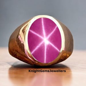 Könnte beinhalten: Ein goldener Ring mit einem großen, ovalen, rosafarbenen Sternrubin. Der Rubin hat ein sternförmiges Muster aus weißen Linien, die vom Zentrum ausstrahlen.