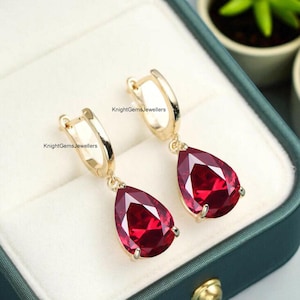 Puede incluir: Un par de pendientes de aro dorados con gemas rojas en forma de lágrima. Los pendientes se muestran en una caja de joyería verde azulado. El texto "KnightGemsJewellers" está impreso en los pendientes.