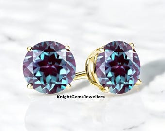 Peut inclure: Une paire de boucles d'oreilles à clous avec des pierres précieuses rondes et facettées à effet changeant, serties dans du métal doré. Les pierres précieuses présentent des nuances de violet, de bleu et de vert. Les boucles d'oreilles sont présentées sur une surface blanche.