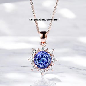 May include: A rose gold necklace with a pendant featuring a large, round, deep blue gemstone surrounded by smaller, clear gemstones. The pendant is starburst-shaped. The necklace is displayed on a white surface.