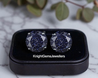 May include: A pair of dark blue gemstone stud earrings with a sparkling, speckled appearance, set in silver. The earrings are displayed in a black jewelry box. The text "KnightGemsJewellers" is visible on the box.