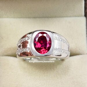 Könnte beinhalten: Ein silberner Ring mit einem großen, oval geschliffenen, tiefroten Edelstein in der Mitte. Die Ringschiene ist breit und poliert und auf beiden Seiten mit zwei Reihen kleiner, klarer, funkelnder Steine besetzt. Der Text KnightGemsJewellers ist sichtbar.