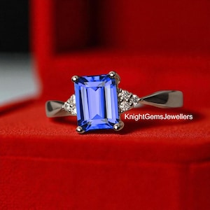 Pode incluir: Um anel de prata com uma grande pedra preciosa azul retangular como peça central. Pequenas pedras transparentes ladeiam a pedra principal de cada lado. O anel está sobre um fundo vermelho, com o texto "KnightGemsJewellers" visível.