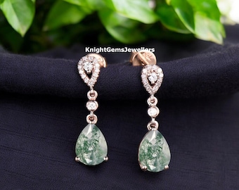 May include: Rose gold drop earrings with a teardrop-shaped moss agate stone. The earrings feature a halo of small diamonds around the top and three smaller diamonds in the middle. The earrings are displayed on a dark blue surface.