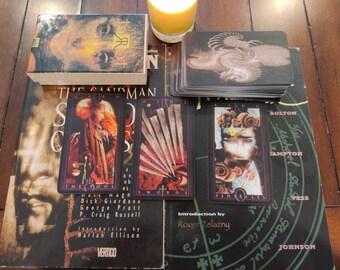 May include: A deck of The Sandman Tarot cards featuring a box with a face illustration, a stack of cards with a swirling design, and three laid-out cards: The Fool, Nine of Swords, and King of Pentacles, resting on a Sandman graphic novel.