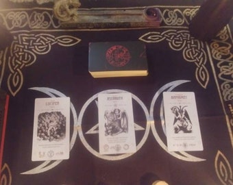 May include: A black cloth featuring a white triple moon and pentagram symbol, displaying three tarot cards labeled Lucifer, Asmodeus, and Baphomet. A deck of cards with a red circular emblem sits above, flanked by two books titled Occult Tarot and Portable Darkness.