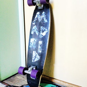 May include: A black skateboard with purple wheels and the word "MARA" written in white letters on the deck. The skateboard is leaning against a wall and is on a blue and green rug with a cartoon monster design.