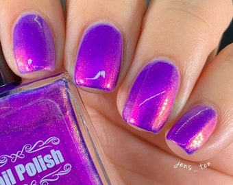 May include: Close-up of a hand with polished nails. The nails are painted a vibrant purple with a color-shifting effect, transitioning to a pinkish-red hue at the tips. A bottle of nail polish is partially visible, with the label reading "ail Polish".