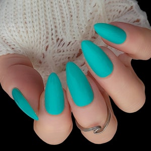 May include: Close-up of a hand with five turquoise almond-shaped nails. The nails are painted a bright turquoise color and have a matte finish.