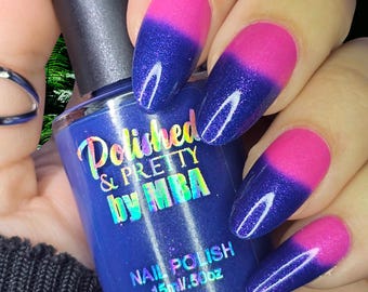 May include: A bottle of blue nail polish with the text "Polished & Pretty by MBA" is held near a hand with nails painted in a pink and blue gradient. The nail polish bottle is 15 ml / .50 oz.