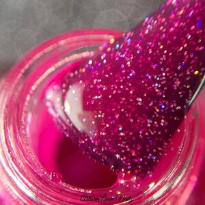 Pu&ograve; includere: Primo piano di un flacone di smalto per unghie rosa con un pennello immerso nello smalto. Lo smalto &egrave; rosa glitterato con glitter iridescenti.