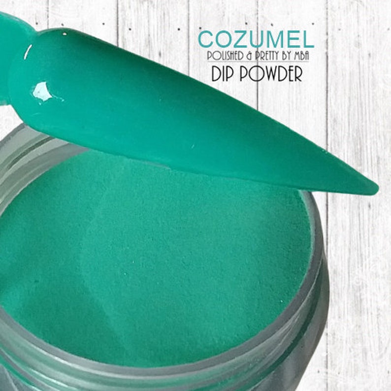 May include: A jar of teal green dip powder for nails. The jar is open and a teal green nail dip brush is resting on the rim of the jar. The text on the jar reads "COZUMEL POLISHED & PRETTY BY MBA DIP POWDER".