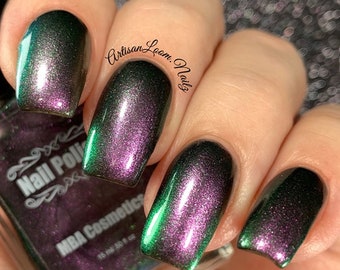 May include: Close-up of fingernails painted with a dark green and purple iridescent nail polish. The polish is from the brand "MBA Cosmetics" and is labeled "Nail Polish".
