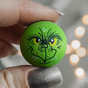 May include: A green painted wooden ball with a hand-painted Grinch face. The Grinch has yellow eyes, a black nose, and a white mouth.