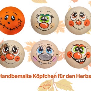 May include: Six hand-painted wooden spheres with cartoon faces, including an orange pumpkin-like sphere. The spheres have various expressions with painted eyes, noses, and mouths. The text "Handbemalte Köpfchen für den Herbst" is at the bottom.