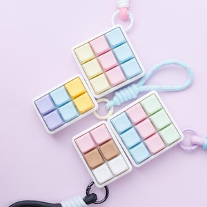May include: Four square-shaped keychains with a grid of colorful pastel blocks. Each keychain has a white frame and a different color scheme, including pink, blue, yellow, and brown. The keychains are attached to pastel-colored lanyards.