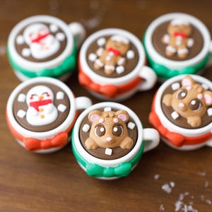 May include: Six festive mugs filled with hot chocolate, each topped with a different holiday-themed design. The designs include a snowman, gingerbread man, and a bear. The mugs are decorated with red, green, and white accents.