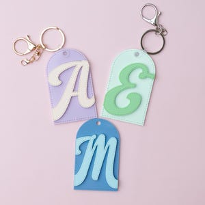 May include: Three keychains with initial letter designs. The keychains feature a light purple, light green, and light blue tag with the letters "A", "E", and "M" in white and light blue. Each tag has a metal keyring and clasp.