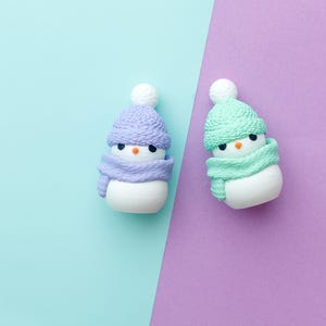 May include: Two snowman figurines, one in lavender and the other in mint green, are set against a split background of light blue and purple. Each snowman wears a matching hat and scarf, with black eyes and an orange nose.