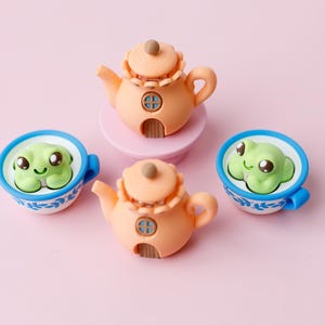 May include: A whimsical arrangement of miniature ceramic teapots and teacups. The peach-colored teapots have decorative details and a small door. The teacups are blue and white, each containing a smiling green frog. The background is a soft pink.