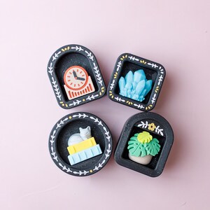 May include: Four small, black decorative trays with white leaf and dot designs. Each tray holds a different miniature item: a clock, a blue crystal, a cat on books, and a potted plant. The background is a solid pink.