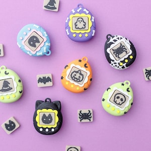 May include: A collection of Halloween-themed digital pet toys in various colors, including blue, purple, orange, black, and green. Each toy features a different spooky icon on its screen, such as a ghost, bat, or spider. Small matching tiles are scattered around.