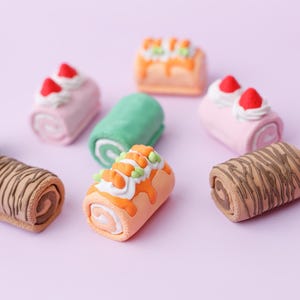 May include: A collection of miniature, colorful Swiss roll cakes on a light purple surface. The cakes feature various designs, including pink with strawberries, green, orange with frosting, and chocolate.