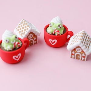 May include: A festive arrangement featuring miniature gingerbread houses and red mugs with heart designs. Each mug holds a whimsical creature with whipped cream and a candy cane. The scene is set against a soft pink background.